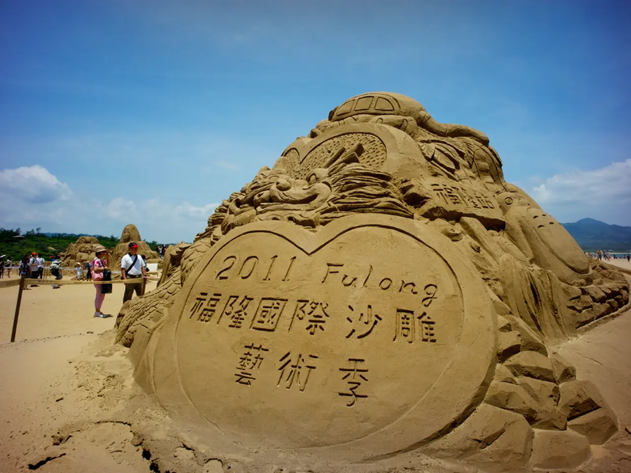 In this image I see the art which is made of sand and I see number of people and in the background...