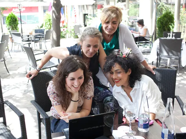 There are four women here looking at the laptop which is on the table. On the table we can see...