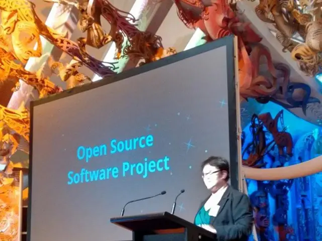 In this image there is a screen, monitor, podium, mics, person and decorative objects. Something is...