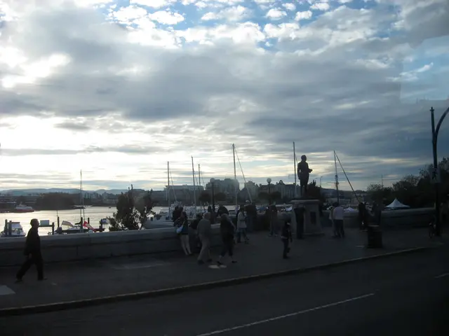 In the image there are many people walking on the road and behind there are ships in the sea and in...